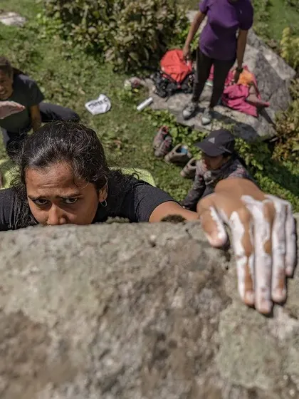 A climber reaches for a hold, her hand covered in chalk. This top-down view shows the commitment and trust in the spotters below.