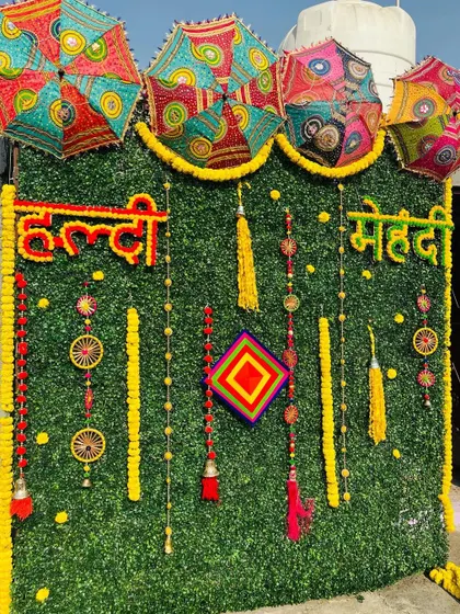 For a modern take on traditional Haldi and Mehndi decor, I use an artificial green wall as a base. This backdrop is decorated with marigold garlands, colorful tassels, and Rajasthani umbrellas to create a vibrant and festive photo corner.