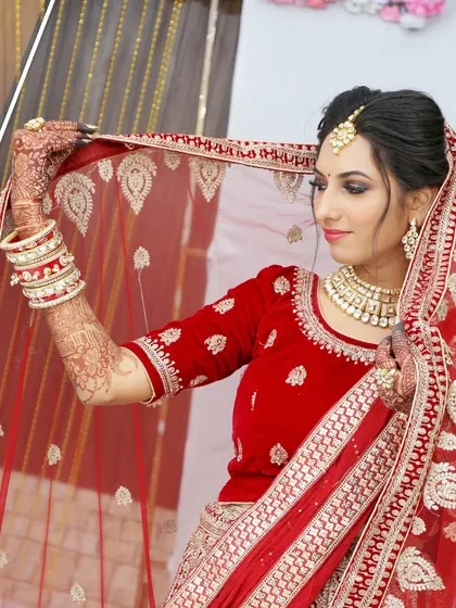 A North Indian bride in a red velvet lehenga, adjusting her dupatta (veil) in a classic pose.