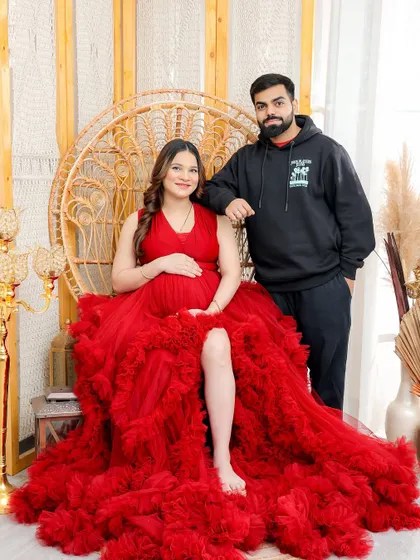 This wide shot shows the full splendor of the red gown and the boho-chic studio setup, creating a beautiful portrait of the expecting couple.