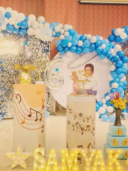 A music-themed 6th birthday party stage. The backdrop includes a blue sequin wall, a blue balloon arch, and custom-printed pedestals with musical notes, all tailored to the 'Samyak' birthday theme.