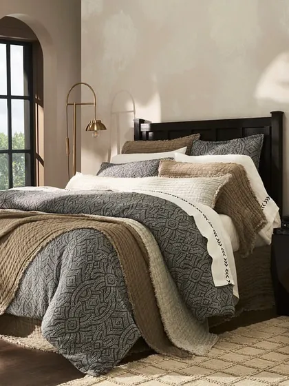 An elegantly styled bed with a mix of textured and patterned linens in a neutral color palette. The arched windows and soft lighting contribute to a sophisticated and serene atmosphere.