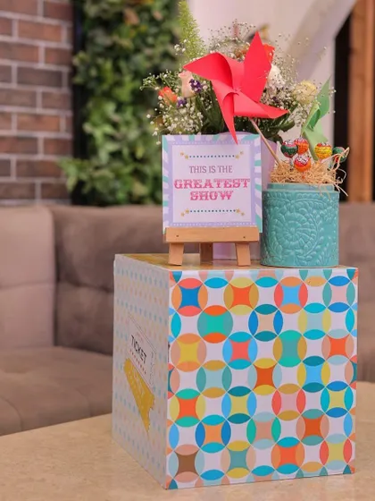A colorful and geometric-patterned box serves as a stand for this carnival centerpiece. The display includes a mini-easel with a "Greatest Show" sign, a red pinwheel, and fresh flowers, adding a playful touch to the table decor.