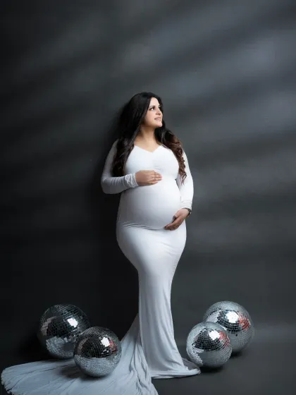 A modern and stylish maternity look. The mother-to-be poses with disco balls, adding a fun and glamorous touch to her classic white gown portrait.