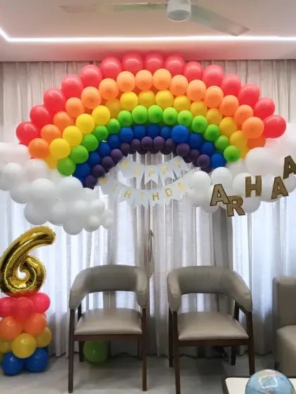 A bright and cheerful rainbow-themed birthday decoration. A large balloon rainbow serves as the main backdrop, complete with fluffy white balloon clouds.