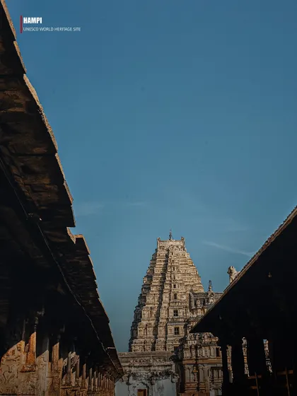 A low-angle shot looking up at the gopuram (tower) of the Virupaksha Temple, framed by the roofs of the surrounding structures. The composition emphasizes the temple's towering height and intricate carvings.