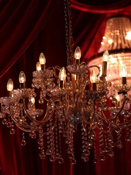 A detailed shot of a classic crystal chandelier against the rich red fabric. The warm light from the bulbs creates a soft, romantic glow, perfect for a wedding reception or engagement party.