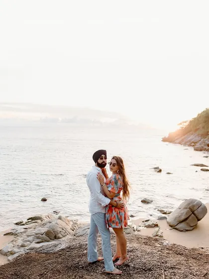 A classic romantic portrait on a rocky shore in Thailand, with the couple embracing against the backdrop of the sea and sky.