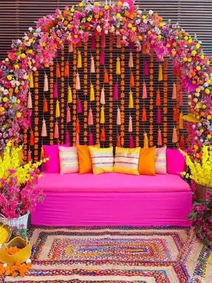 A stunning Mehendi seating area with a dense floral arch of pink and yellow flowers. The backdrop features hanging tassels, and the space is decorated with colorful cushions and a woven rug.