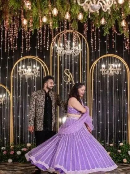 A beautiful couple's portrait from a Sangeet or reception. The backdrop features golden arches, hanging chandeliers, and a curtain of fairy lights, creating a dreamy effect.