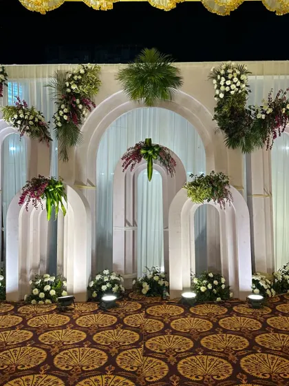 An elegant and contemporary stage backdrop featuring a series of white arches of varying sizes. The structure is tastefully decorated with white and burgundy floral arrangements.