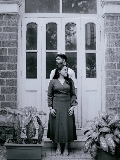 A classic black and white portrait of a couple standing in front of a large, traditional door. The composition is symmetrical and elegant, creating a timeless feel.