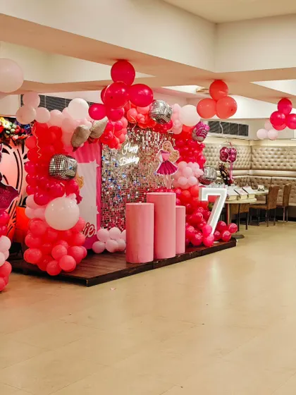 A large-scale 7th birthday party in a banquet hall. The stage is decorated in shades of pink and red with a shimmer wall and multiple props.