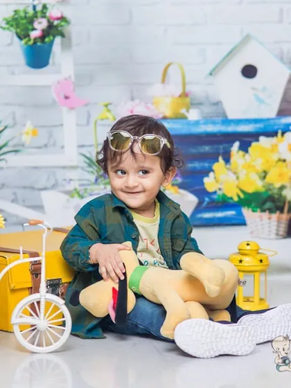 A cool little dude in sunglasses, sitting amongst a colorful spring-themed setup with yellow flowers, birdhouses, and a miniature bicycle. His relaxed pose is full of personality.