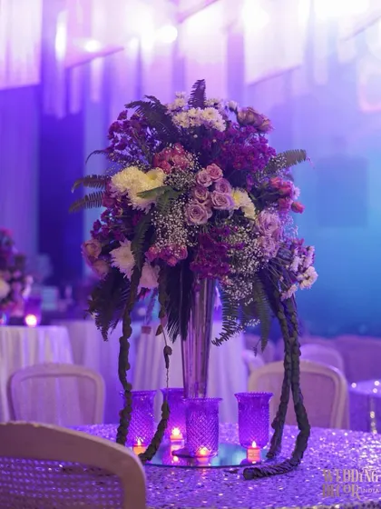A tall floral centerpiece on a table with a sequined purple cloth. The dramatic uplighting creates a glamorous and celebratory atmosphere.