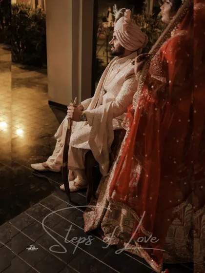 An artistic, candid shot of the couple. The focus on the groom, seated with his kirpan, while the bride stands beside him, creates a powerful and stylish image.