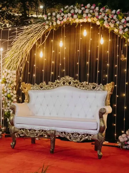 A romantic photo booth or seating area with a classic white and gold sofa. The circular arch is adorned with flowers and fairy lights, creating a perfect spot for memorable pictures.