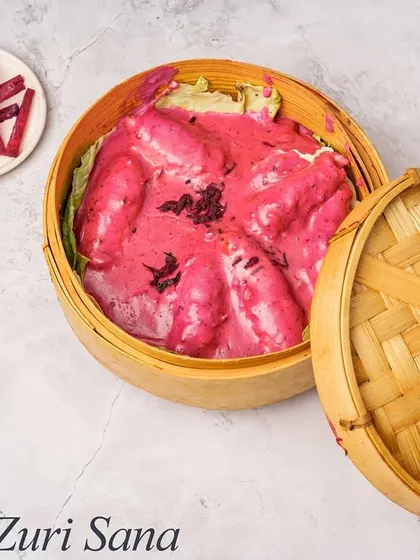 Zuri Sana Chicken is a unique dish featuring tender chicken in a vibrant, creamy beetroot-based sauce. It is served in a traditional bamboo steamer, which keeps it warm and adds to the presentation.