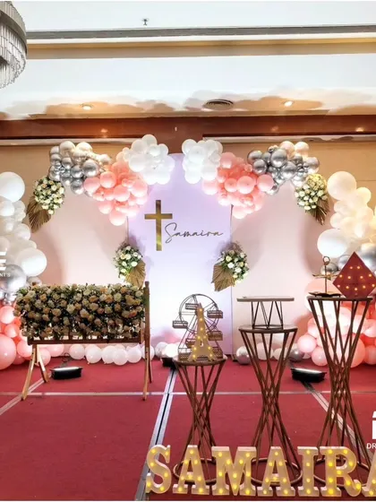 A lovely Baptism party for Samaira, featuring a soft pastel palette of pink, white, and silver. The balloon arch frames a personalized backdrop with a cross motif, and marquee lights spell out her name.