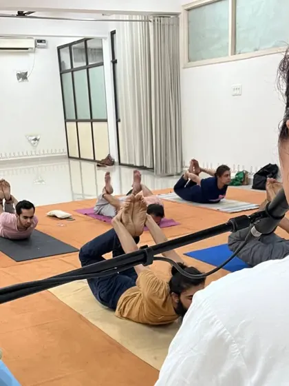 A teacher on the dais guides a full class through various asanas, ensuring each participant practices correctly and safely.