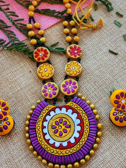 A large, round mandala-style pendant necklace with floral motifs in purple and yellow. The matching jhumkas complete this bold and beautiful set.