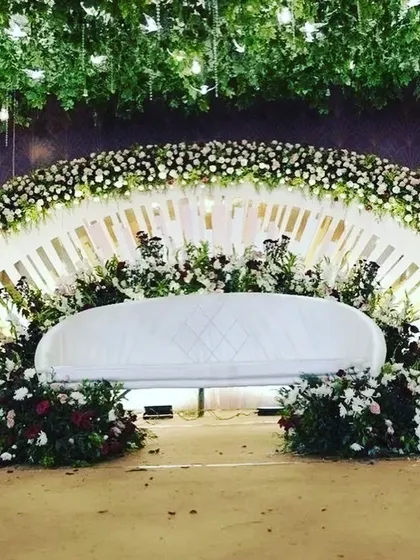 A beautiful white-themed stage with a fanned backdrop and a ceiling of hanging greenery and birds. The lush floral arrangements in white and deep red create a classic and romantic look.