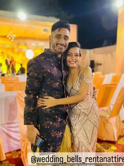 Fun and stylish outfits for a sangeet or reception. The bride is wearing a glamorous golden gown with tassels, and the groom is in a dark floral print Indo western jacket.