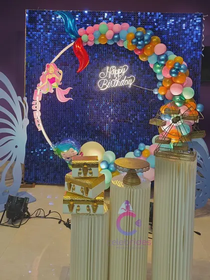 A magical mermaid and butterfly-themed birthday setup. The blue sequin backdrop, colorful balloon arch, and butterfly wing props create a fantasy world.