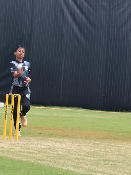 A young player delivers the ball during a match, with the umpire watching closely.