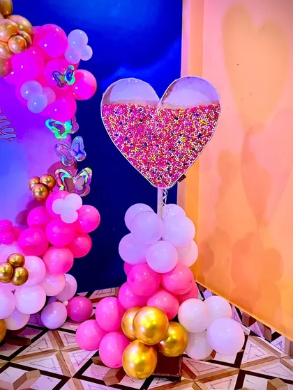A unique decorative piece for a naming ceremony. This heart-shaped stand is filled with colorful sprinkles and surrounded by pink, white, and gold balloons.