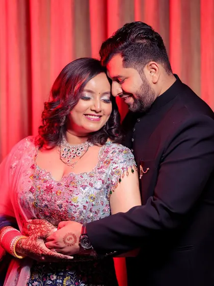 This is a warm, romantic portrait from an engagement celebration. The couple holds each other in a gentle embrace, their smiles showing their comfort and love. The red curtain backdrop adds a touch of drama and richness to the scene.