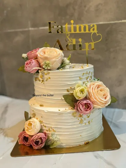 A classic two tier engagement cake with a textured buttercream finish. It's adorned with soft pastel roses and a sprinkle of gold leaf, perfect for celebrating Fatima and Adin's special day.