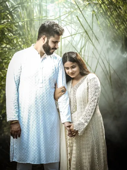 A quiet, intimate moment during a pre-wedding shoot. The couple is dressed in traditional white outfits, creating a soft and romantic mood amidst a natural, green setting.