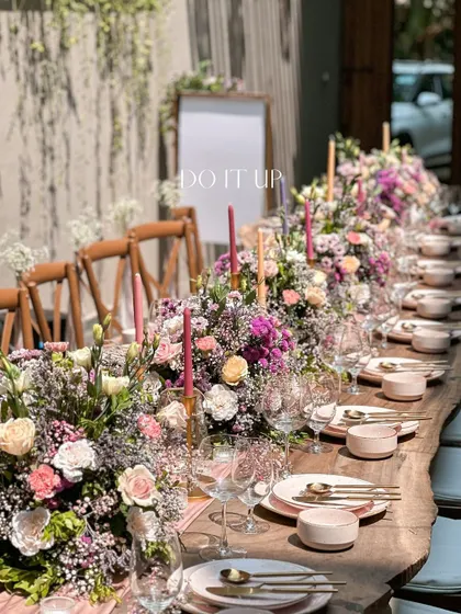 A long-table setup for an intimate bridal shower. The wooden table is adorned with a dense runner of pink and purple flowers, creating a lush and luxurious feel for the celebration.