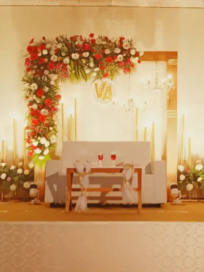 A romantic and warm stage setting perfect for an anniversary or reception. The design uses an L-shaped floral arrangement of red and white roses, complemented by numerous candles for soft lighting.