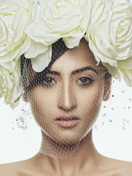 A portrait titled "Cloistered," featuring a model behind a delicate veil and a crown of white roses. The image plays with themes of confinement and beauty, creating a soft yet thought-provoking editorial visual.