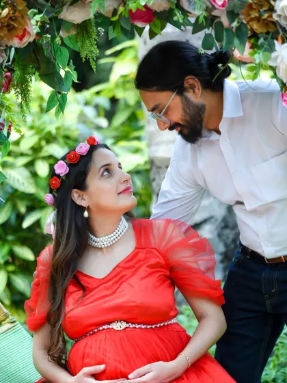 A beautiful, intimate portrait of an expecting couple. The mom-to-be, wearing a red dress and a floral headband, shares a loving look with her partner.