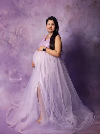 A full-length shot showcasing the beautiful drape of the lavender tulle maternity gown. This classic pose highlights the baby bump and the elegance of the expecting mother.
