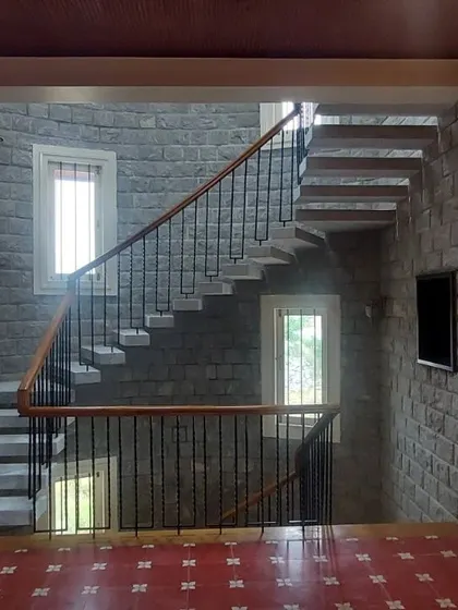 This interior showcases a symphony of natural materials. A floating staircase with wrought iron railings ascends against a backdrop of solid stone walls, while the red Kota stone flooring adds warmth and traditional character.