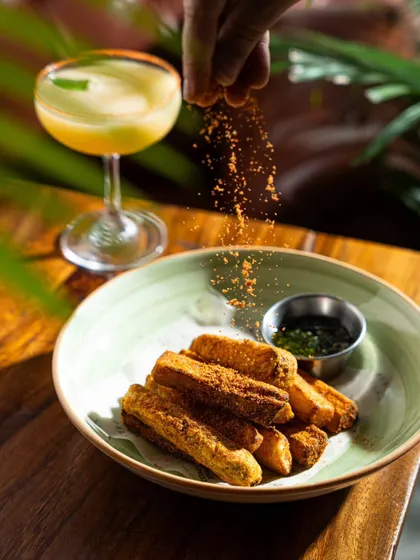Another angle of seasoning being sprinkled on fries, with a cocktail in the background. This composition connects the food and drink experience.