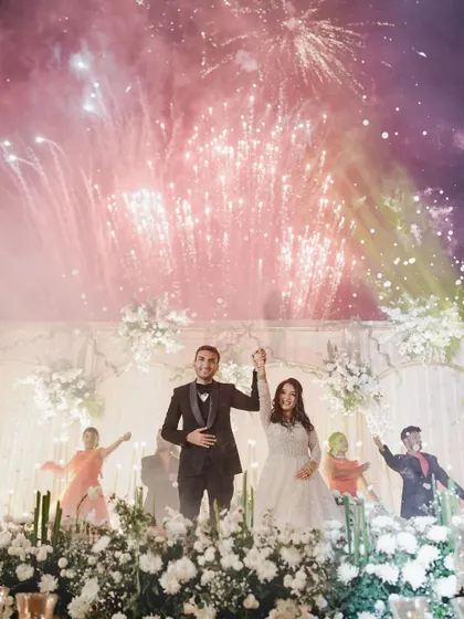 A spectacular moment as fireworks light up the sky behind the couple on stage. This image perfectly captures the grand scale and celebratory spirit of a luxury wedding Sangeet.