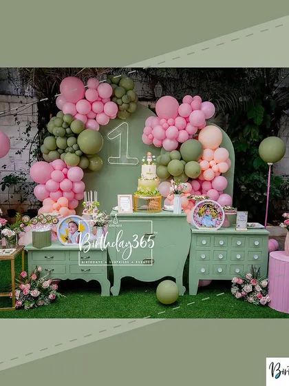 An alternate angle of the stylish sage green and pink first birthday decor. This view shows the cake table, custom photo displays, and the overall arrangement of props and balloons on a turf-grass floor.