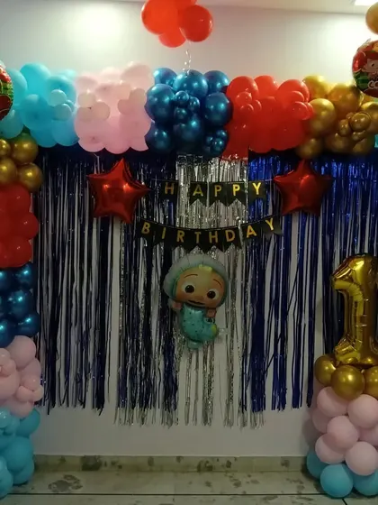 A colorful Cocomelon party setup for a first birthday. This decoration features a blue tinsel curtain, a multi-colored balloon frame, character foil balloons, and a large number '1' foil balloon.