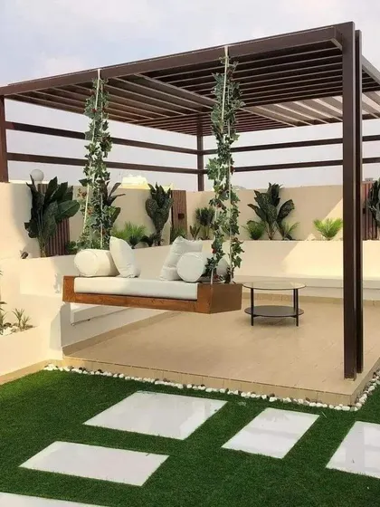 A beautiful rooftop lounge with a hanging swing sofa under a large pergola. The artificial grass and white stepping stones add a garden-like feel to the terrace.