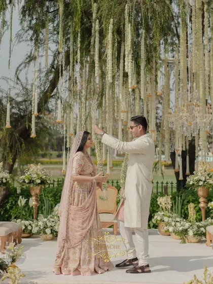 The Jaimala ceremony begins. The groom playfully bows as the bride places the garland, a moment of joy captured against the beautiful backdrop of hanging florals.