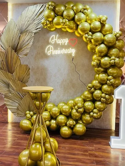 A chic all-gold anniversary decoration. The setup includes a balloon ring, a "Happy Anniversary" neon sign, a light-up number "1", and gold palm leaf accents.