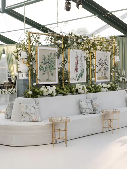This elegant lounge seating features a custom backdrop with botanical prints, framed in gold and adorned with fresh white flowers and greenery. It serves as both a seating area and a beautiful photo opportunity.
