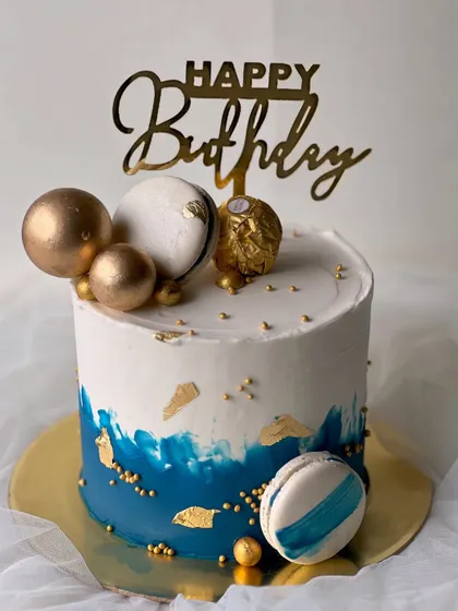 A stylish and masculine design perfect for him. This rich vanilla buttercream cake features a blue watercolor effect, gold leaf, macarons, and a Ferrero Rocher topper.