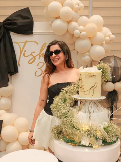 The birthday girl posing with her stunning cake, which is displayed on a custom stand adorned with baby's breath. The black and white bow theme is carried through from the backdrop to the cake itself.