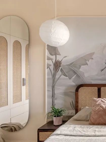 A serene bedroom view where a large oval mirror reflects the custom cane-detailed wardrobe, expanding the sense of space. The nature-inspired mural and paper lantern pendant add layers of softness and tranquility.